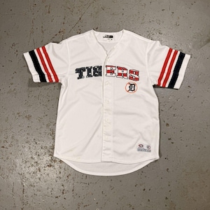 Op de afbeelding: Wit honkbalshirt met het woord "TIGERS" over de borst in zwarte en rode letters. De mouwen hebben rood-zwarte strepen. Een "D"-logo staat op de rechterborst.