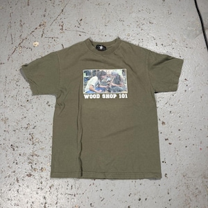 May include: Olive green t-shirt with a rectangular graphic featuring two people working with wood. The text "WOOD SHOP 101" is printed below the graphic. The shirt has short sleeves and a crew neck.