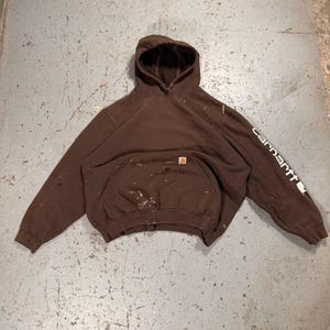 May include: A brown Carhartt hoodie with a hood and a front pocket. The word "Carhartt" is printed in white on the left sleeve. The hoodie has paint splatters on the front.