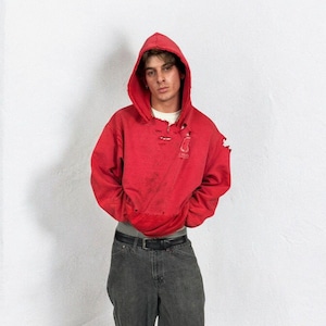 May include: A distressed red hoodie with a drawstring hood and a small logo. The hoodie is worn with grey jeans and a black belt. The outfit has a vintage, worn appearance.