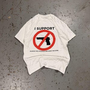 Puede incluir: Camiseta blanca con el texto "I SUPPORT" sobre un círculo rojo con una pistola negra tachada. Debajo del círculo, el texto dice "SILENCE THE VIOLENCE AND SHUN THE GUNS". La camiseta está sobre una superficie de hormigón gris.