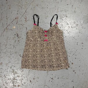 Può includere: Un top a camisole con stampa leopardata. Presenta spalline sottili nere con dettagli rosa e fiocchi rosa al centro. Il tessuto è beige con macchie marroni. Un capo elegante.