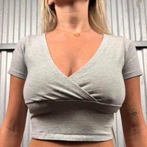 May include: A light gray, short-sleeved crop top with a V-neckline. The top features a wrap-style design across the chest. The fabric appears to be a soft, knit material. The top is a solid color and has a simple, modern design.