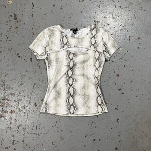May include: A short-sleeved top with a snakeskin pattern in shades of grey and white. The top features a keyhole cutout detail below the neckline. The garment is made of a soft, form-fitting material.