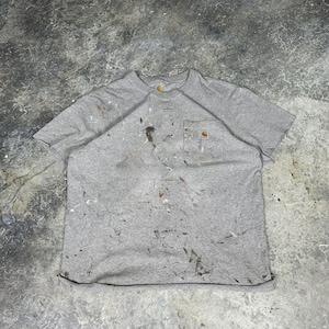 May include: A grey Carhartt t-shirt with a chest pocket, covered in paint splatters. The shirt is displayed flat, revealing the crew neck and short sleeves. The Carhartt logo is visible on the pocket.