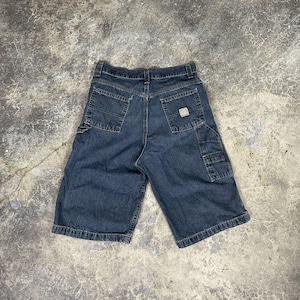 Puede incluir: Pantalones cortos vaqueros azul oscuro con múltiples bolsillos, incluido un bolsillo lateral utilitario. Los pantalones cortos tienen un diseño clásico de cinco bolsillos y un pequeño parche con el logotipo en la parte posterior. Los pantalones cortos están planos sobre una superficie gris texturizada.