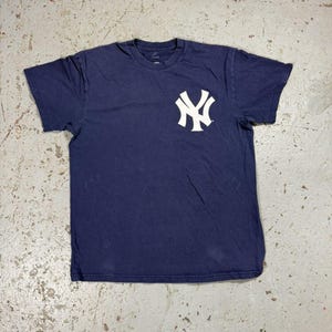 May include: Navy blue t-shirt featuring the New York Yankees logo in white on the chest. The shirt is a classic crew neck style, made of a soft, likely cotton material. The shirt is laid flat on a concrete surface.