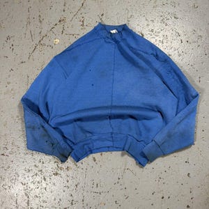 Puede incluir: Una sudadera azul vintage con cuello alto y mangas largas. La prenda muestra signos de desgaste, con manchas oscuras en las mangas y el cuerpo. La sudadera está tendida sobre una superficie gris.