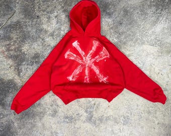 May include: A red hooded sweatshirt with a white, abstract design on the front. The sweatshirt has long sleeves and a cropped fit. The design appears to be hand-painted or screen-printed.