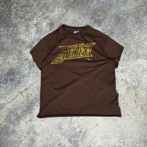 Puede incluir: Una camiseta marrón con un gráfico dorado de la palabra "JOM" en una fuente estilizada.