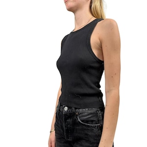 Può includere: Un top senza maniche nero a coste. Il top senza maniche ha una scollatura alta e una silhouette aderente. Il tessuto sembra essere un materiale morbido ed elastico. Il top senza maniche è abbinato a jeans neri.