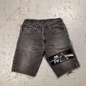Pode incluir: Calções de ganga pretos com um aspeto desgastado. Um patch dos Pink Floyd é costurado na perna direita. O patch apresenta um fundo preto com um triângulo branco, o nome da banda e um arco-íris.