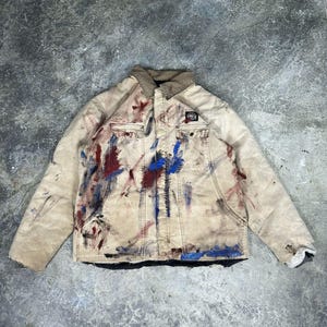 May include: A tan work jacket with a brown collar and multiple pockets. The jacket is splattered with red, blue, and black paint. A small patch is visible on the right chest pocket.