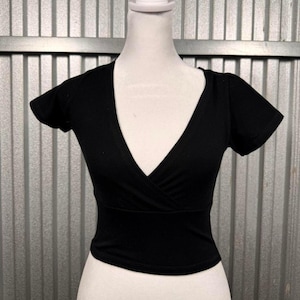 May include: A black wrap-style top with short sleeves and a deep V-neck. The top is displayed on a white mannequin, showcasing its fitted silhouette and design. The fabric appears to be a soft, solid color.