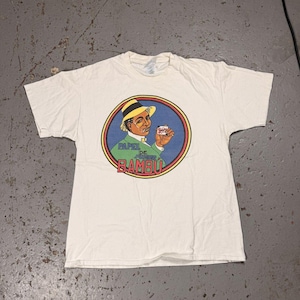 Könnte beinhalten: Cremefarbenes T-Shirt mit einem kreisförmigen Grafikdesign, das einen Mann mit gelbem Hut zeigt, der ein kleines Päckchen hält. Die Grafik wird von einem blauen und gelben Rand umrahmt, mit den Worten "PAPEL DE BAMBU" in Grün.