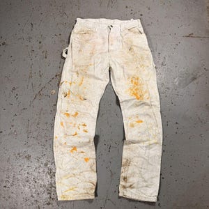 Puede incluir: Pantalones de trabajo blanquecinos con salpicaduras de pintura naranja y marrón. Los pantalones tienen un diseño clásico de cinco bolsillos y cierre de botón. La tela parece desgastada y manchada, lo que sugiere su uso en un entorno de pintura o construcción.