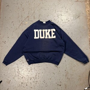 Pode incluir: Camisola azul marinho com a palavra "DUKE" em letras brancas no peito. A camisola é de gola redonda e mangas compridas. A peça de roupa está estendida numa superfície cinzenta.