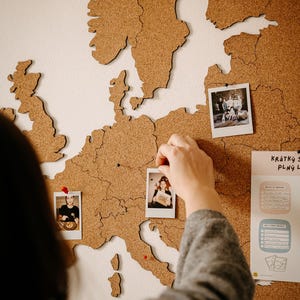 May include: A cork world map of Europe fixed to a white wall, with cut-out continents. Small photos are pinned to the map, and a person is seen placing a photo on the map. The map is a travel-themed decorative piece.