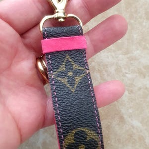 May include: A black and brown patterned keychain with a gold-colored clasp and a bright pink accent. The keychain features a repeating logo pattern and pink stitching. The clasp is a lobster claw style.