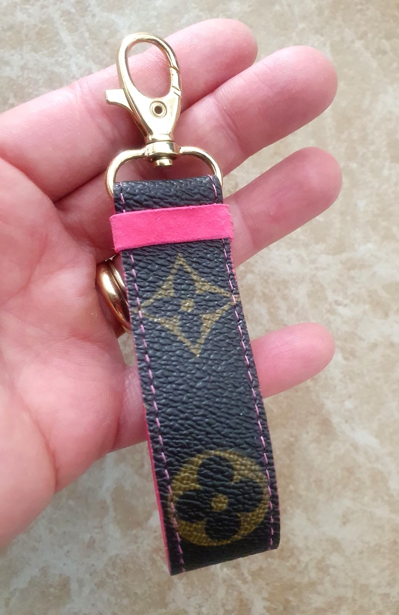 May include: A black and brown patterned keychain with a gold-colored clasp and a bright pink accent. The keychain features a repeating logo pattern and pink stitching. The clasp is a lobster claw style.
