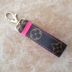 May include: A keychain strap with a gold-colored clasp and a dark brown strap featuring a repeating floral pattern in a lighter brown. A bright pink accent is stitched into the strap.