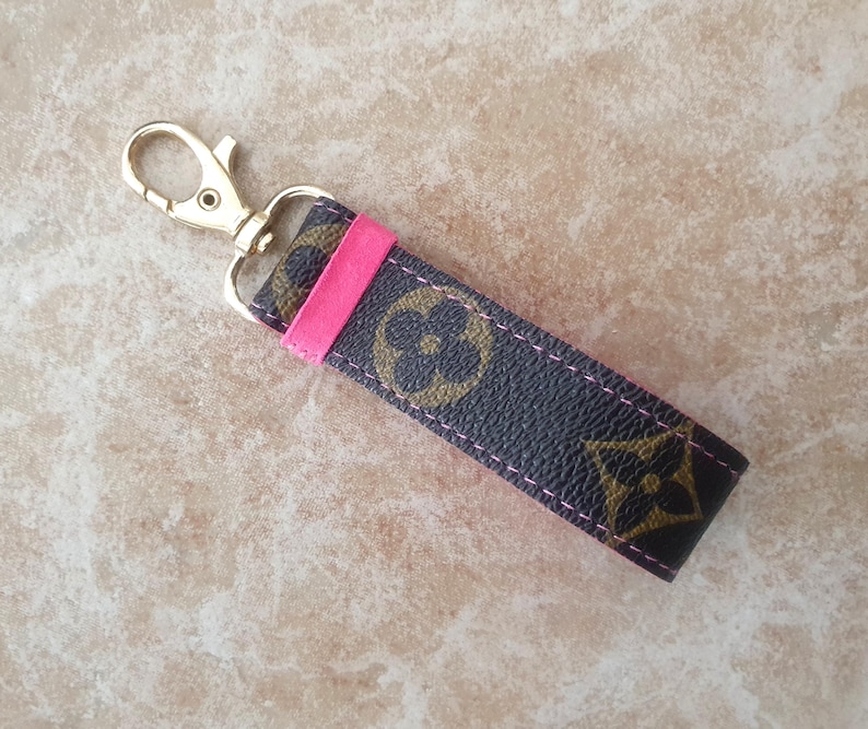 May include: A keychain strap with a gold-colored clasp and a dark brown strap featuring a repeating floral pattern in a lighter brown. A bright pink accent is stitched into the strap.