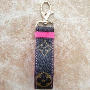 May include: A keychain with a gold-colored clasp and a strap featuring a dark brown pattern with gold-colored floral designs. The strap has a pink accent and pink stitching.