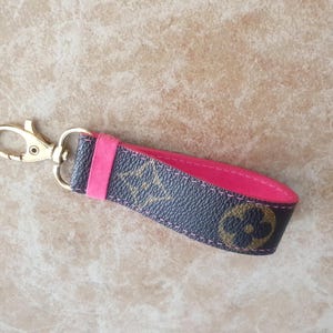 May include: A key fob with a gold-colored clasp and a dark brown and pink strap. The strap features a repeating pattern of gold-colored floral designs. The interior of the strap is a bright pink color.