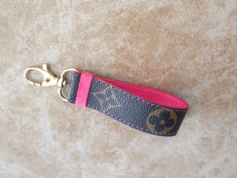May include: A key fob with a gold-colored clasp and a dark brown and pink strap. The strap features a repeating pattern of gold-colored floral designs. The interior of the strap is a bright pink color.