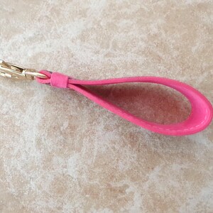 May include: A bright pink leather wrist strap with a gold-colored clasp. The strap is looped and appears to be made of a soft, suede-like material. The clasp is a standard swivel type, suitable for attaching to a bag or other accessory.