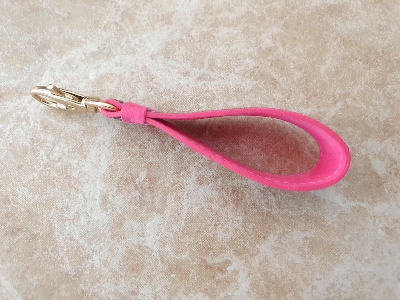 May include: A bright pink leather wrist strap with a gold-colored clasp. The strap is looped and appears to be made of a soft, suede-like material. The clasp is a standard swivel type, suitable for attaching to a bag or other accessory.