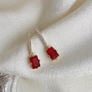 May include: A pair of gold earrings with a red square-shaped stone and a line of clear stones on the dangle.