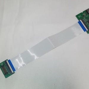 Puede incluir: Una placa de circuito verde con un cable de cinta blanco conectado. La placa de circuito tiene un conector negro en un extremo y un conector azul en el otro extremo.