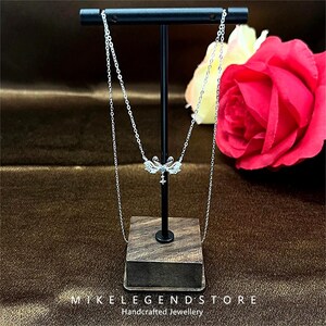 Peut inclure: Un collier en argent avec un pendentif délicat en forme de libellule orné de pierres de zircone cubique transparentes. Le collier est présenté sur un support en bois avec une base noire et un dessus brun. Le texte "MIKE LEGEND STORE Bijoux artisanaux" est visible en bas de l'image.