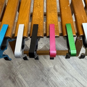 Op de afbeelding: Een verzameling van zes gitaarcapo's in verschillende kleuren: blauw, zilver, zwart, roze, groen en wit. De capo's zijn gemaakt van metaal en zijn ontworpen om op de gitaarhals te klemmen. Ze zijn geplaatst op een houten oppervlak.