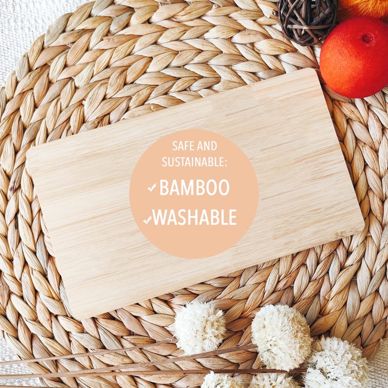 May include: A light brown bamboo cutting board with a round, peach-colored sticker that says "Safe and Sustainable: Bamboo Washable". The board is sitting on a woven straw mat.