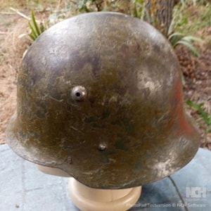 Può includere: Un elmetto da combattimento in metallo verde oliva usurato con forma a cupola arrotondata, tesa svasata e un prominente perno di ventilazione sul lato. La superficie mostra segni di ossidazione, usura e scolorimento su tutto il guscio esterno.
