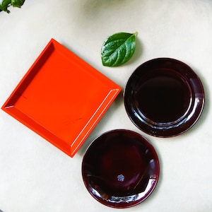 May include: An overhead shot of a bright orange square tray and two dark red round plates. A green leaf is placed near the plates. The plates have a glossy finish, and one has a small floral design.