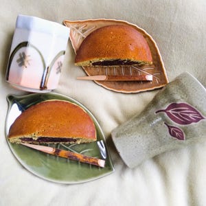 Könnte beinhalten: Draufsicht auf japanische Dorayaki-Pfannkuchen, halbiert, um die rote Bohnenfüllung zu zeigen, serviert auf blattförmigen Tellern mit Holzspießen. Zwei Keramikbecher mit Blumen- und Blättermustern vervollständigen die Szene.