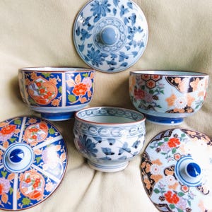 May include: A selection of Japanese ceramic bowls and lids. The bowls display detailed floral and geometric patterns in blue, orange, and white. The lids have a central knob and complement the bowl designs. The set is arranged on a neutral backdrop.