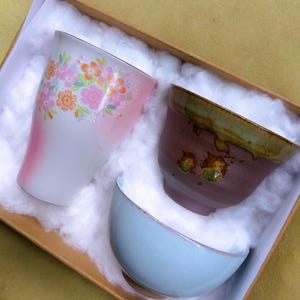 May include: A gift set of three ceramic teacups and bowls. The tall cup is white with pink and orange floral designs. The other cup is brown with a green rim. The bowl is light blue. The set is in a cardboard box with white filler.
