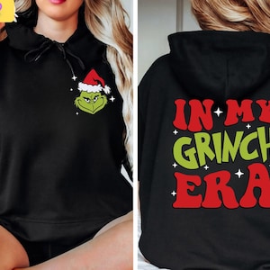 May include: Black hoodie featuring a Grinch graphic on the front and the phrase "IN MY GRINCH ERA" on the back. The Grinch is wearing a Santa hat. The text on the back is in red and green with white stars.