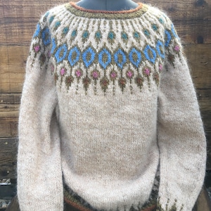 Puede incluir: Un suéter beige de manga larga con cuello redondo. El canesú presenta un patrón geométrico en azul, marrón y rosa. Los puños y el dobladillo están ribeteados con verde y marrón. El suéter se exhibe en un maniquí.