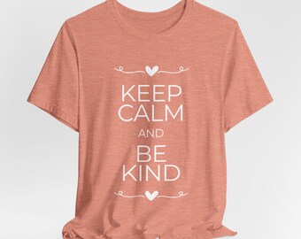 May include: A coral-colored t-shirt with the words "KEEP CALM AND BE KIND" in white, centered on the front. The text is framed by decorative swirls and hearts. The shirt has short sleeves and a crew neck.