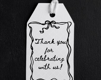 May include: White gift tag with a black border and a string. The tag has the text "Thank you for celebrating with us!" in a handwritten style font. The background is black.