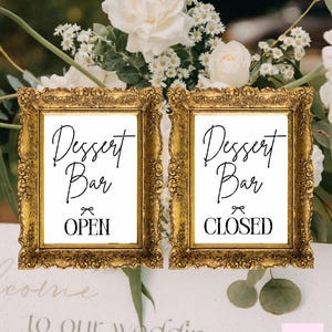 May include: Two gold-framed signs with the words "Dessert Bar" and "Open" and "Closed" in a stylish font. The signs are set against a backdrop of white flowers and greenery, suitable for a wedding.