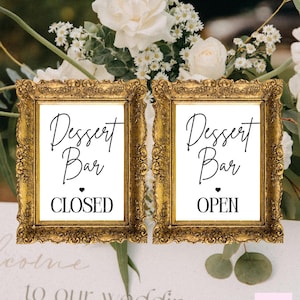 May include: Two framed signs with gold ornate frames. The signs read "Dessert Bar" with a heart symbol, and the words "CLOSED" and "OPEN". The signs are set against a backdrop of white flowers and greenery.