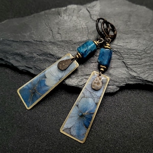Op de afbeelding: Hangende oorbellen met rechthoekige hangers met blauwe bloemmotieven. De oorbellen hebben bronskleurige frames en accenten, met blauwe edelsteen kralen en druppelvormige bedels. Ze worden gepresenteerd op een donkergrijze, getextureerde ondergrond.