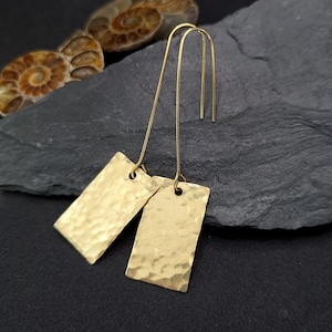 May include: A pair of gold-toned earrings featuring rectangular pendants with a hammered, textured surface. Each pendant is suspended from a long, thin, curved wire hook that passes through a small hole at the top of the metal rectangle.