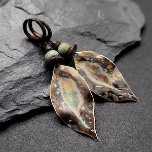 May include: A pair of handcrafted earrings featuring leaf-shaped pendants with a distressed, metallic finish. The leaves display shades of bronze, teal, and gold. The earrings have a dark brown, looped top with small, round, green beads.
